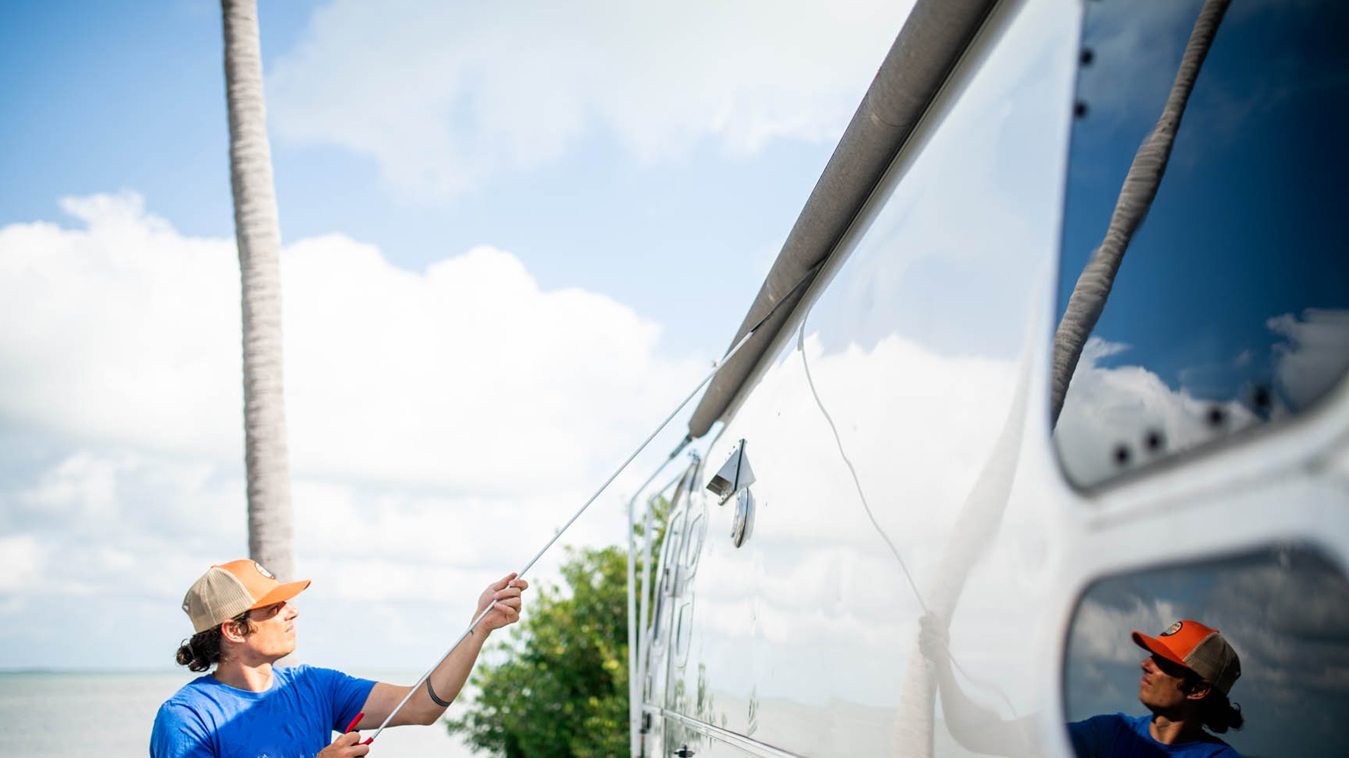 A man pulling out the ZipDee awning on his Airstream travel trailer while parked at a tropical camp site