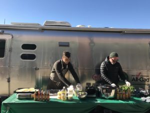 2 men preparing food in front of an Airstream