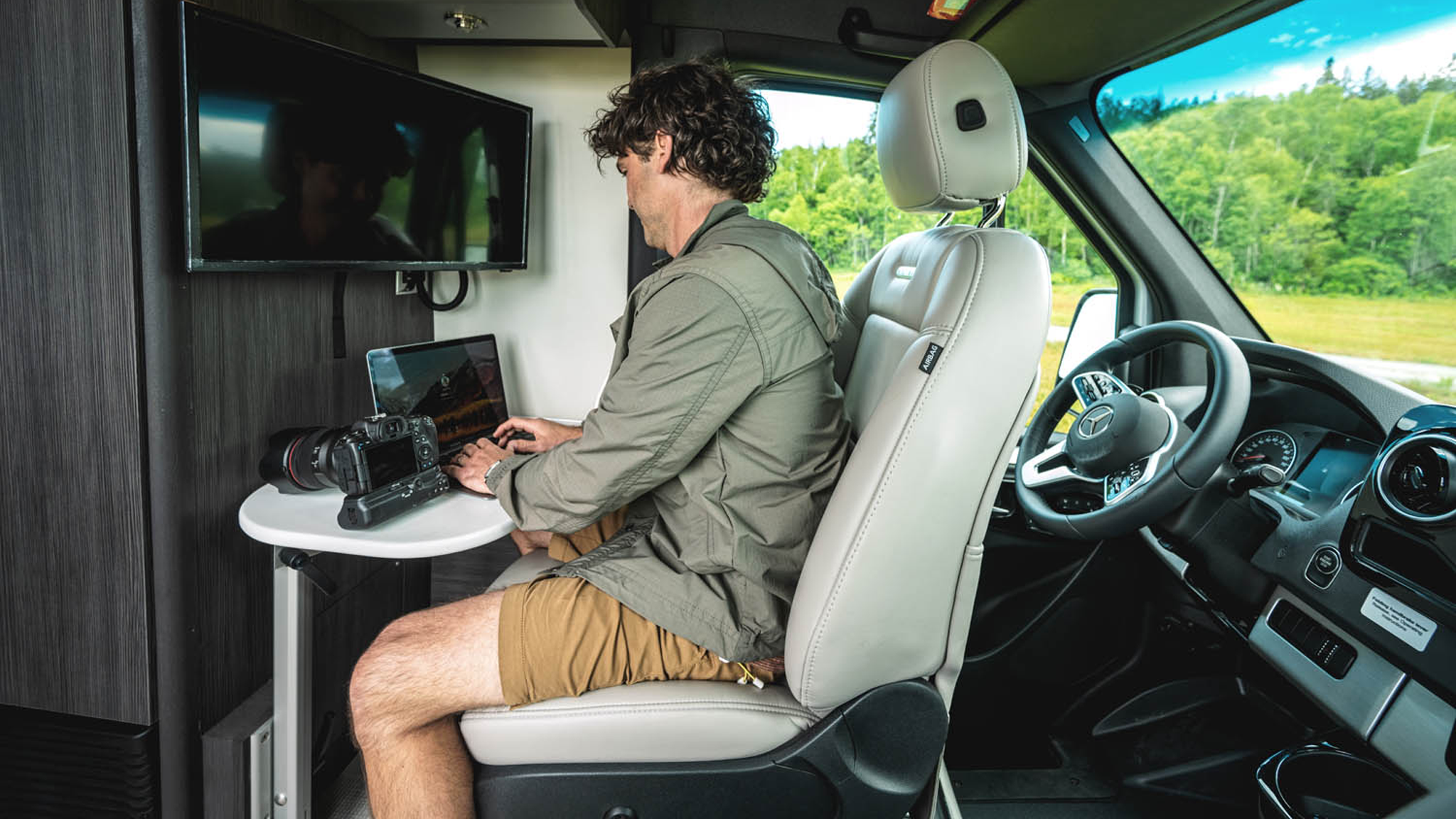 A man working on his computer with his camera sitting on the table inside of an Airstream Interstate Touring Coach