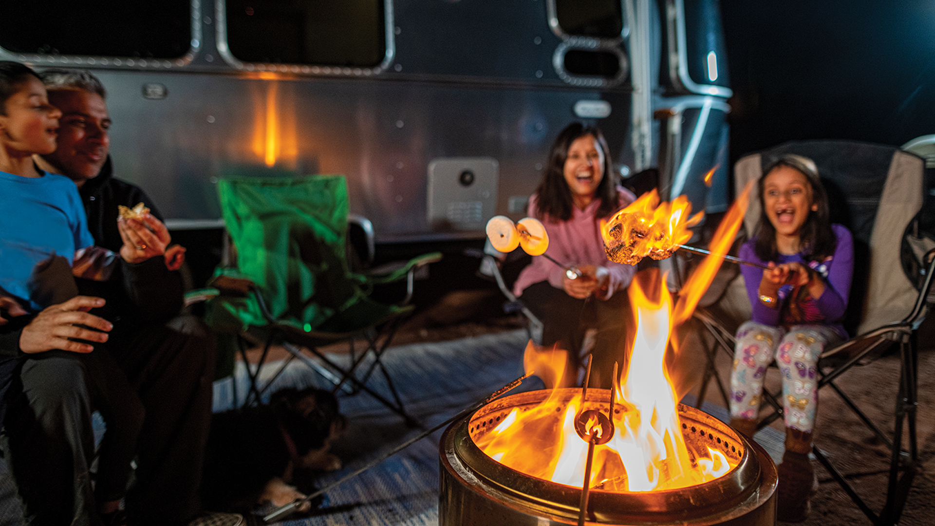 A family sitting in lawn chairs around a camp fire with their Airstream travel trailer behind them