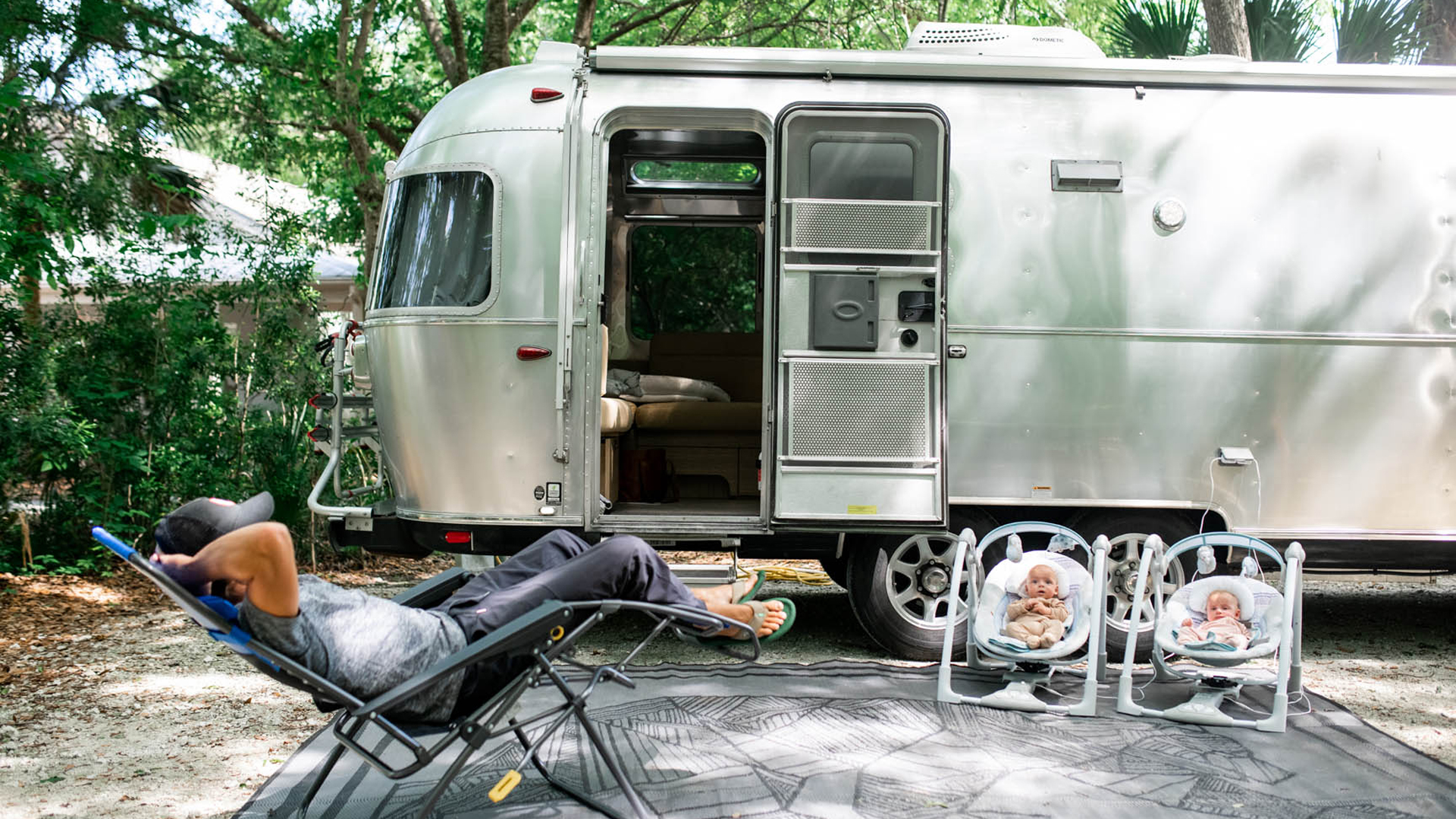 A dad relaxing in lawn chair with twins in baby seats outside of their Airstream travel trailer