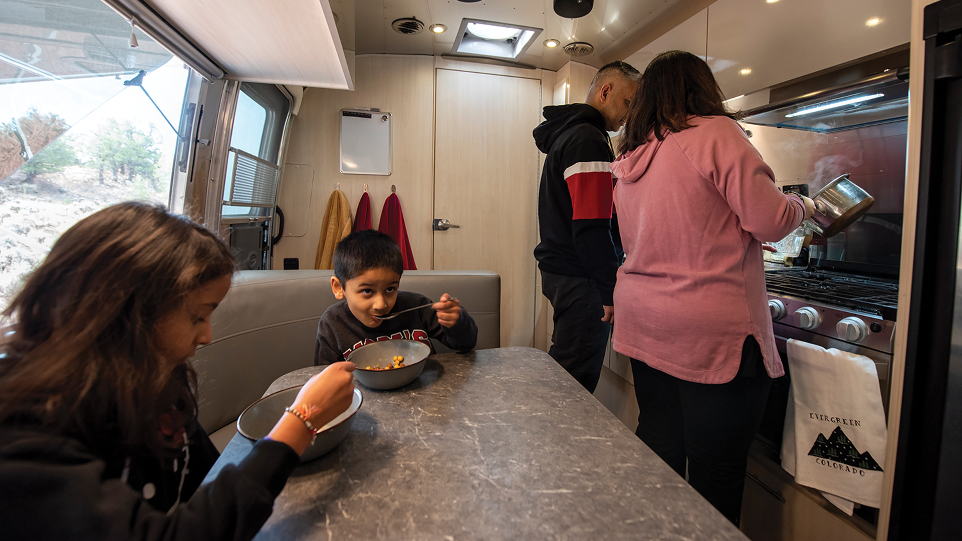 Two kids sitting at a dinette table in an Airstream travel trailer while their parents are cleaning dishes in the kitchen