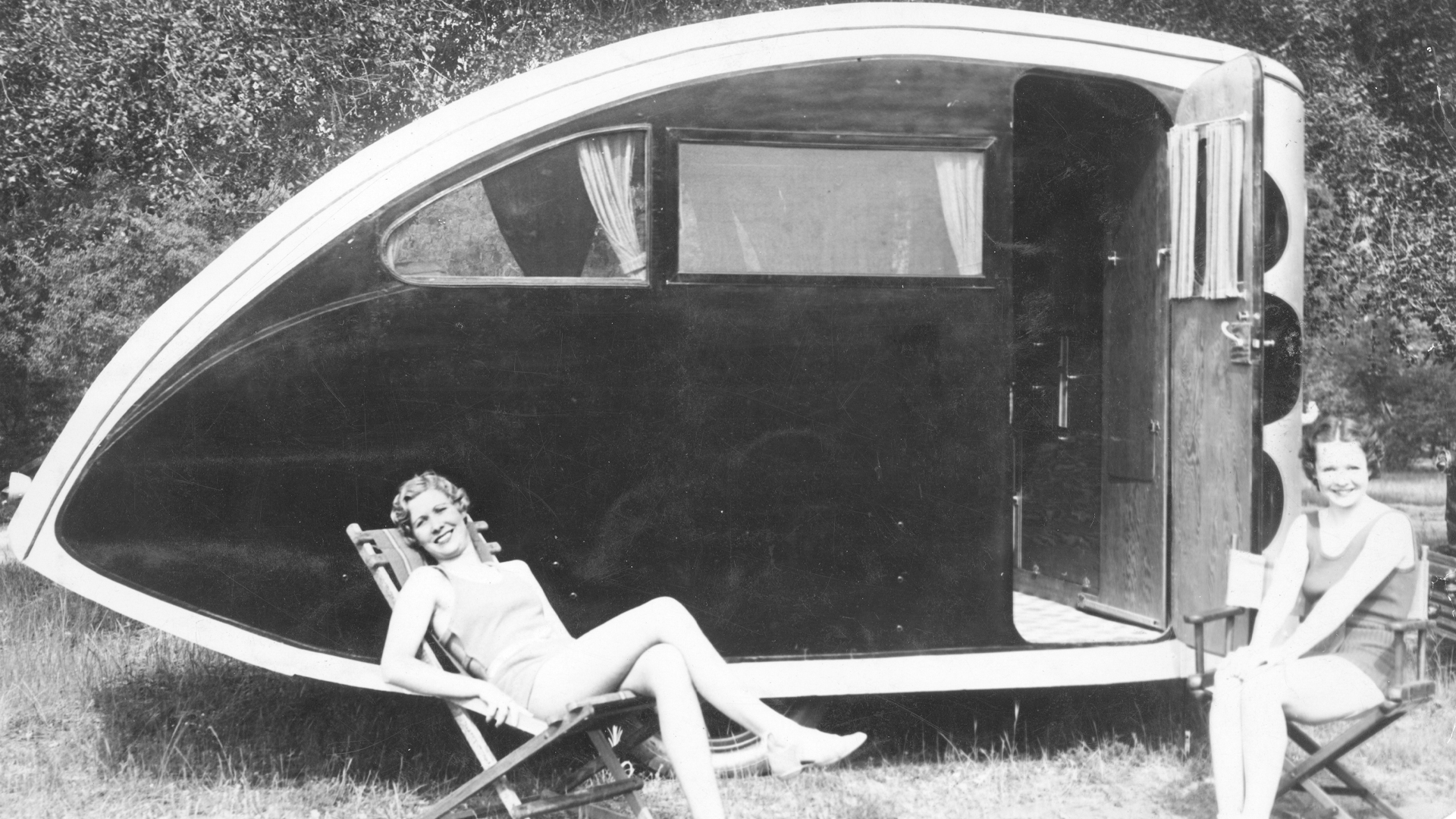 Two women sitting in lawn chairs next to an Airstream Torpedo travel trailer