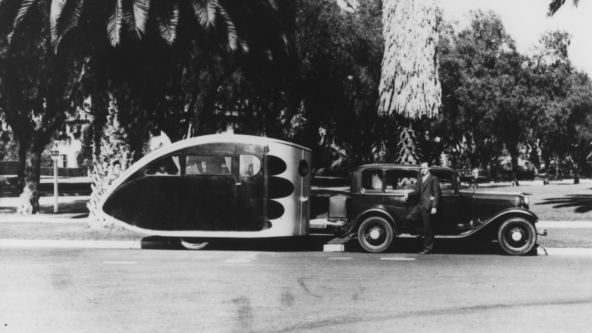An Airstream Torpedo travel trailer being towed by a vintage car in a tropical area