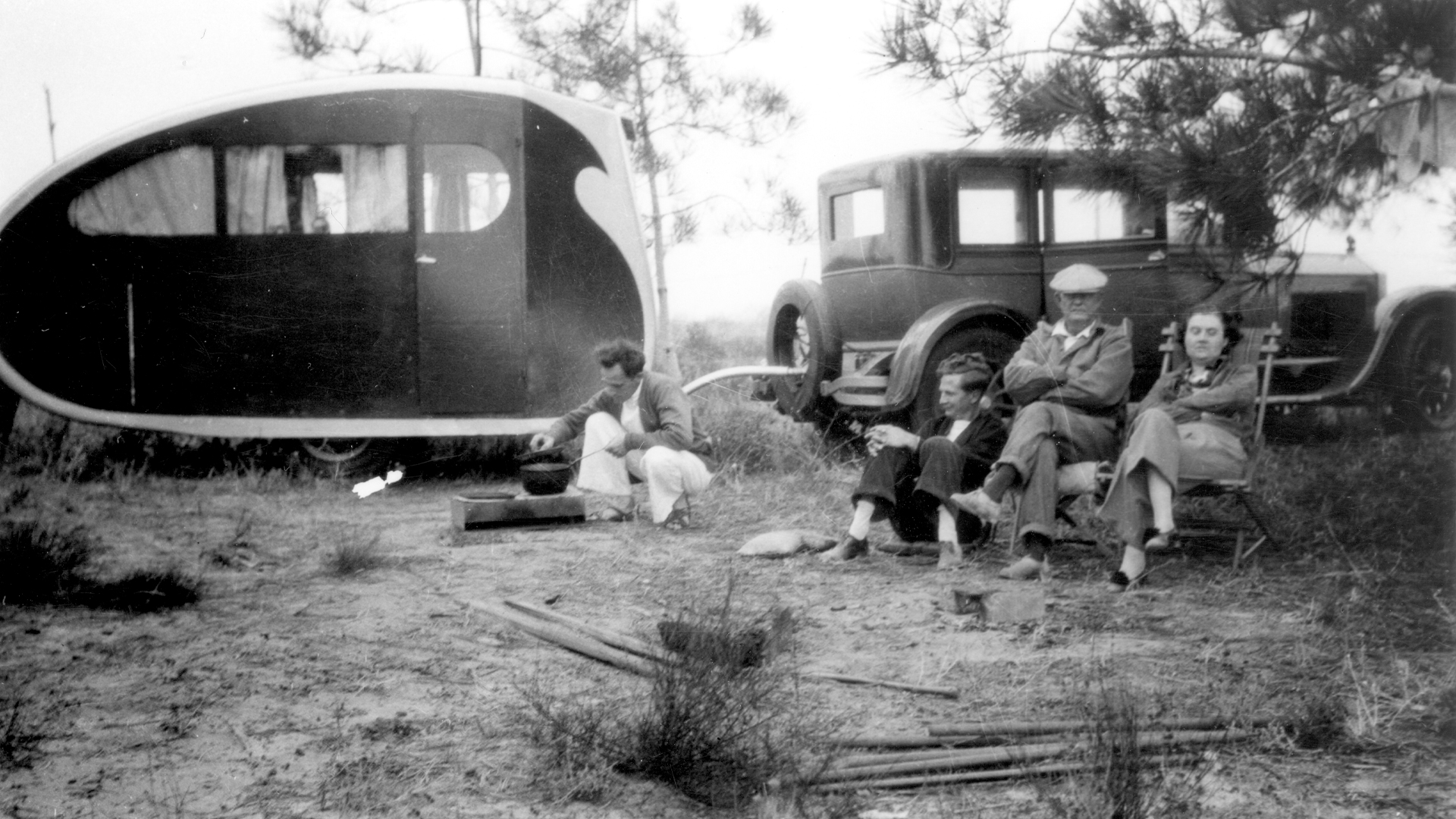 A vintage Airstream Torpedo travel trailer with people sitting outside of the trailer and car.