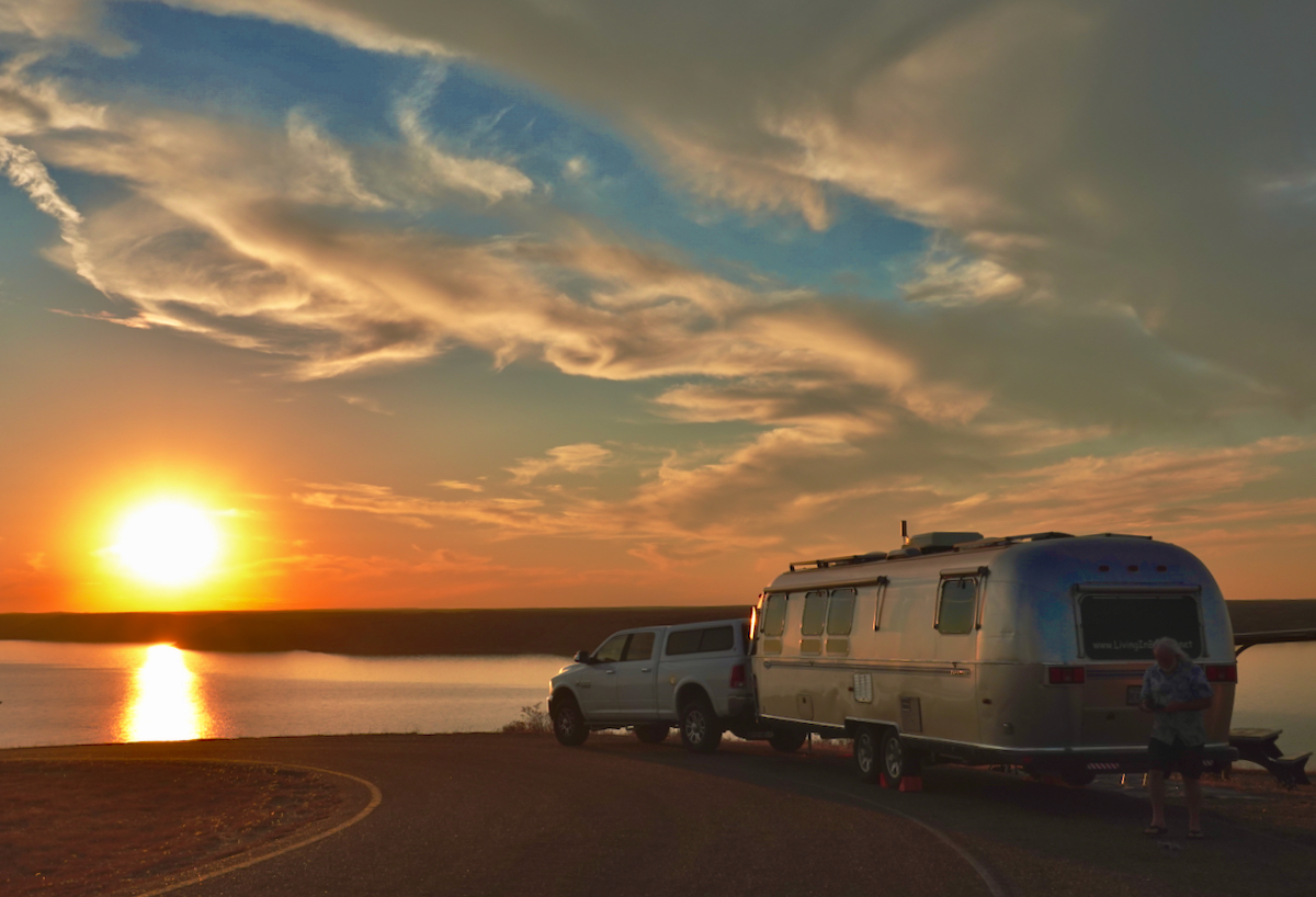 The sun setting over a lake with an Airstream Classic travel trailer and truck parked next to it.