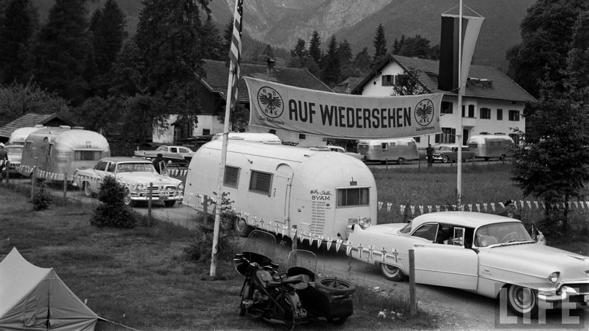 Vintage white trailer Airstream being towed in a white car by Airstream founder, Wally Byam and his wife Stella, as they travel through Europe.