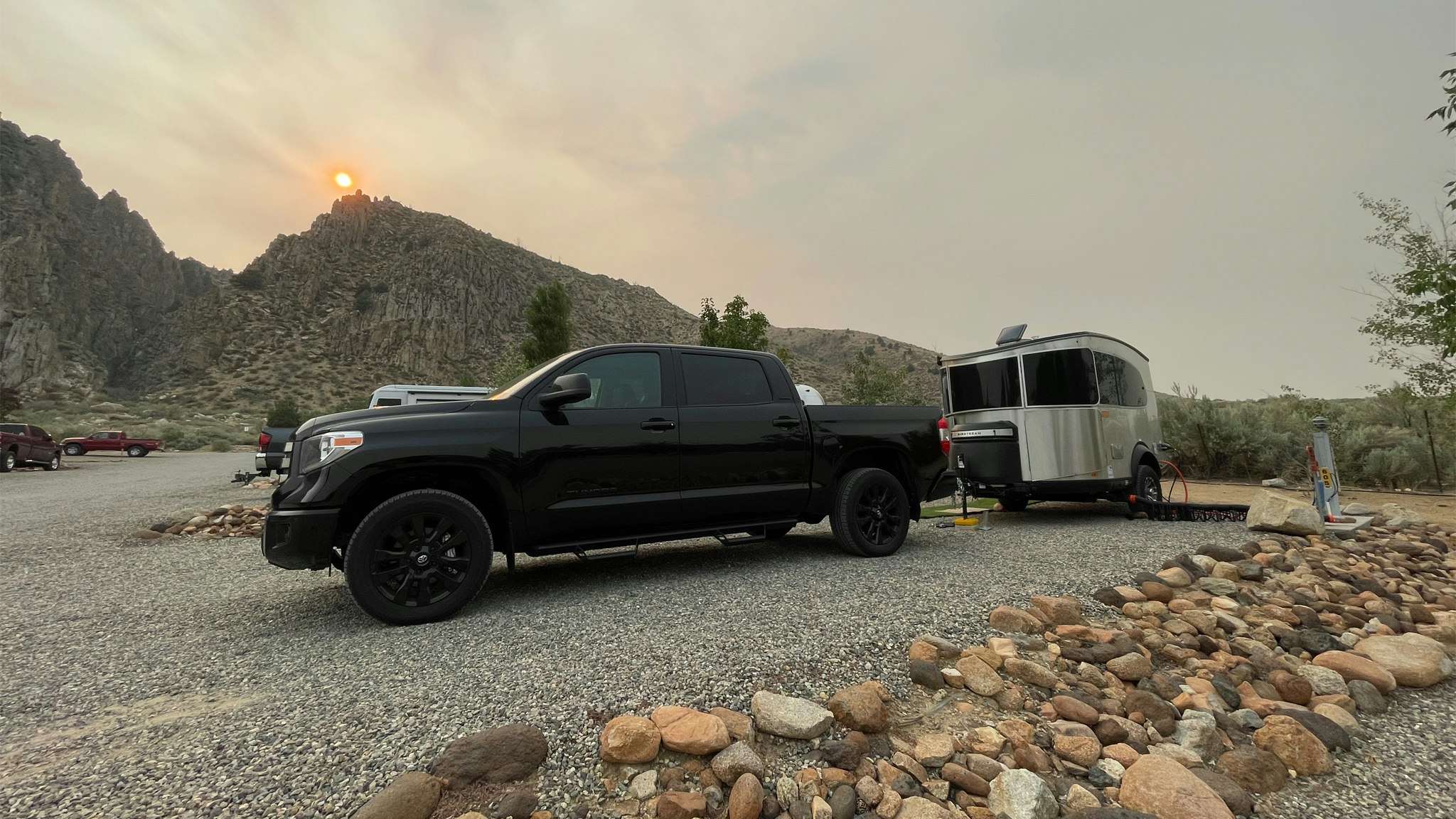 Black truck parking an Airstream Basecamp travel trailer at a campground with mountains and a sunset in the background