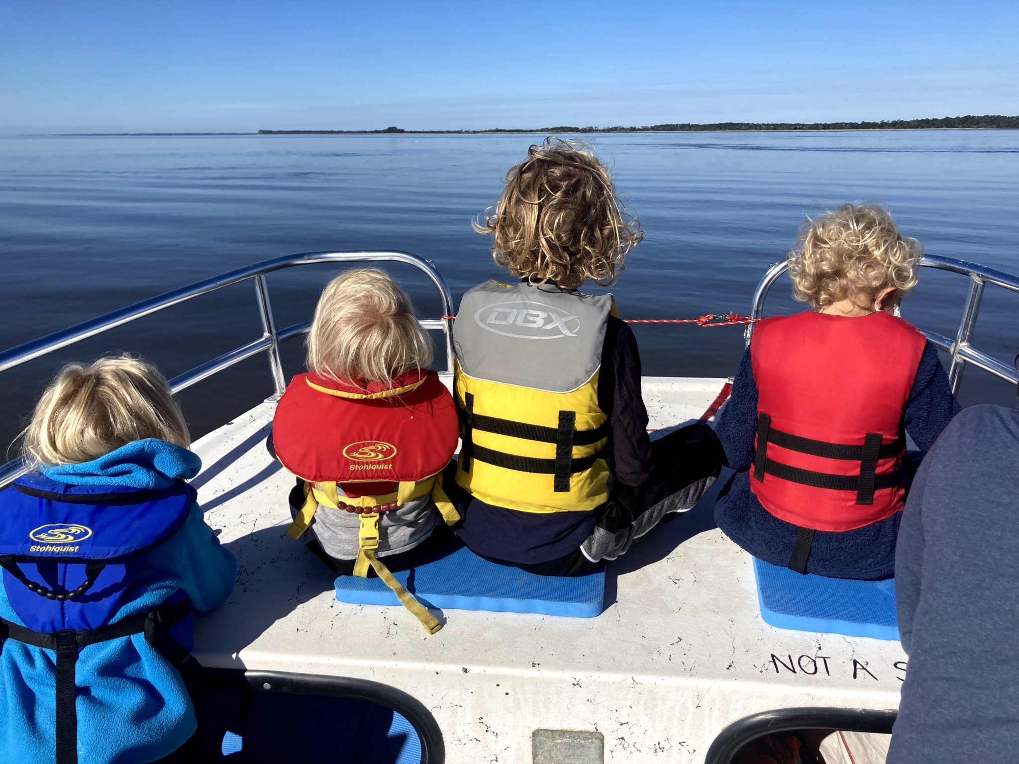 Four kids sitting on the edge of a boat with lifejackets on looking out at the water.