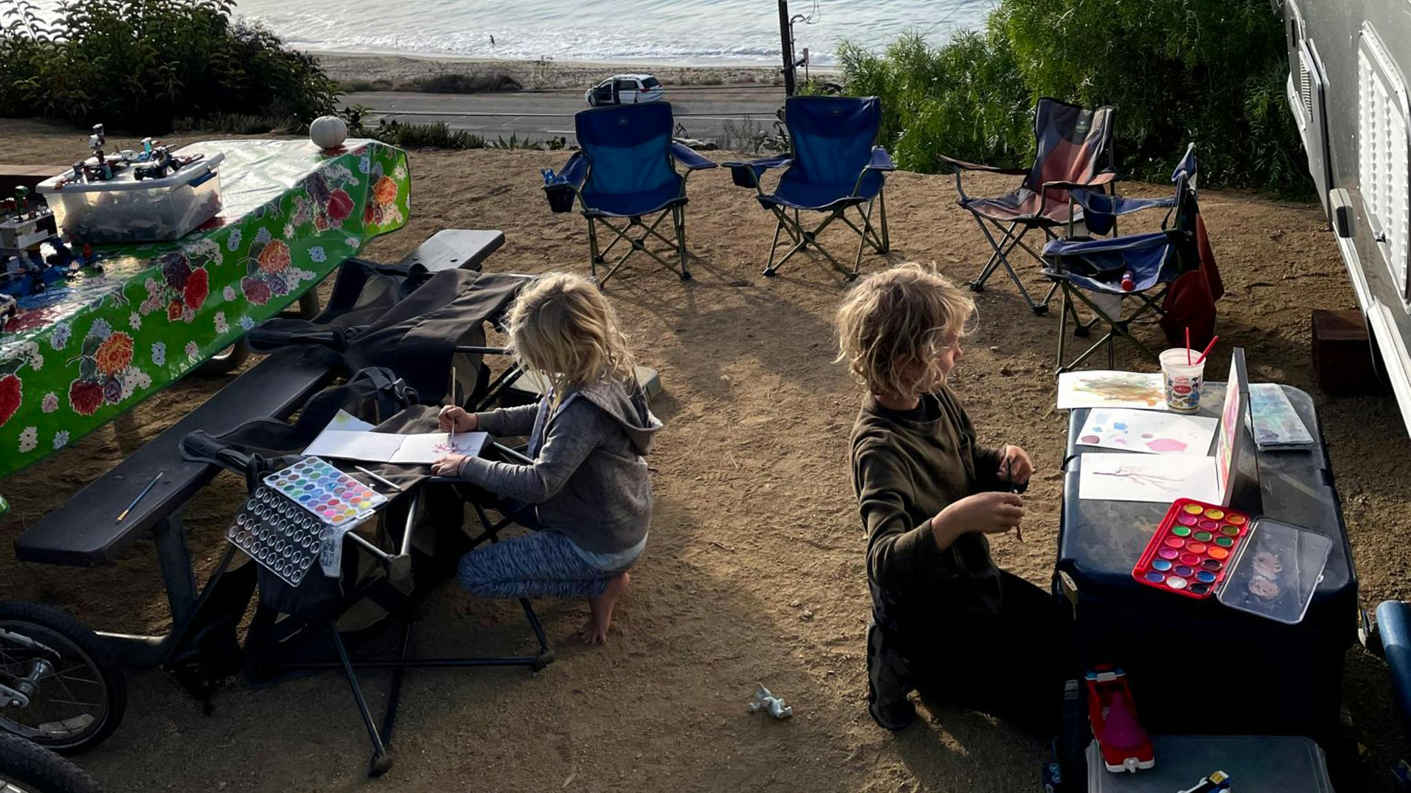 Two kids sitting outside of their Airstream travel trailer at their own tables while they paint next to the water.