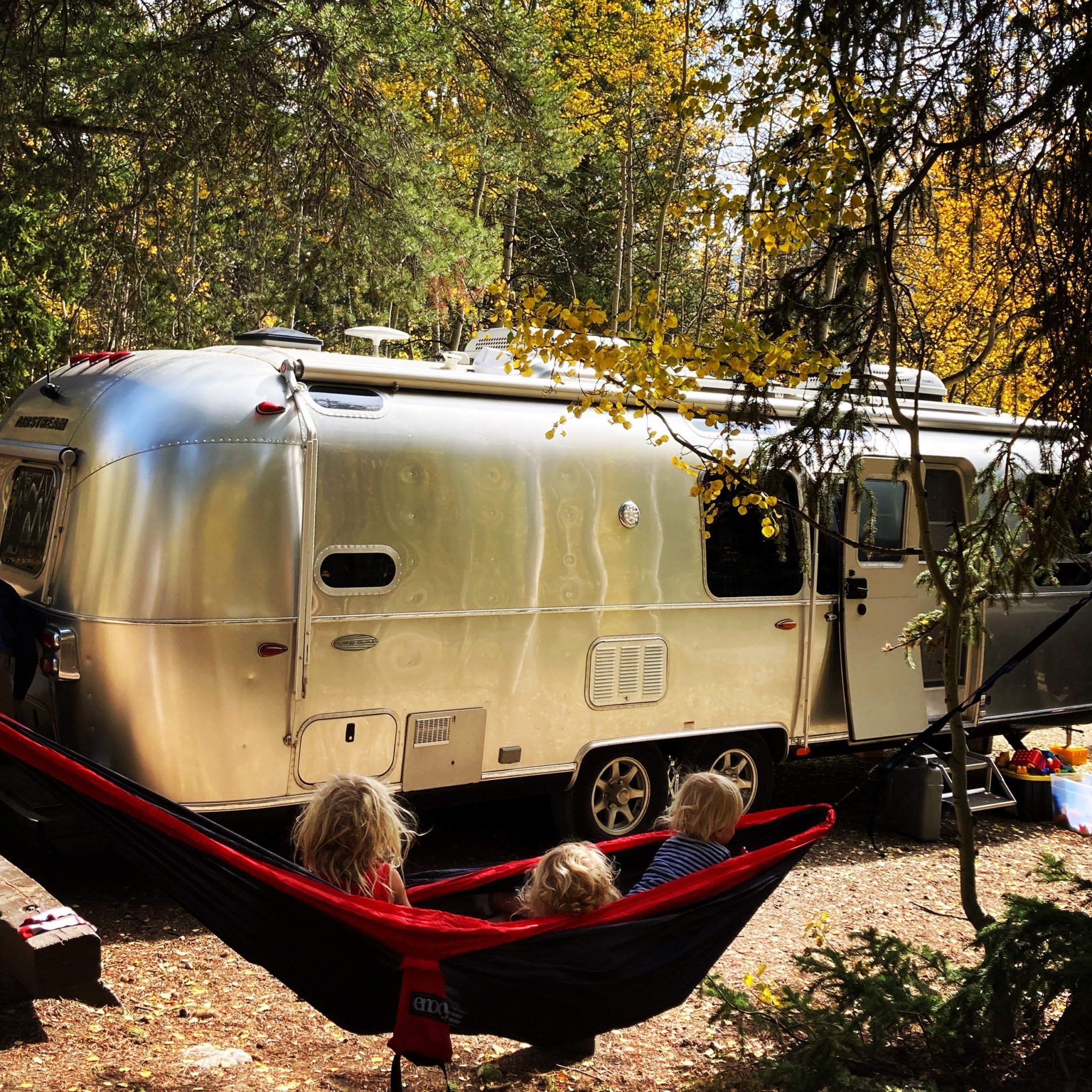 Three kids sitting in a red hammock looking at their Airstream travel trailer that is parked in a woods.