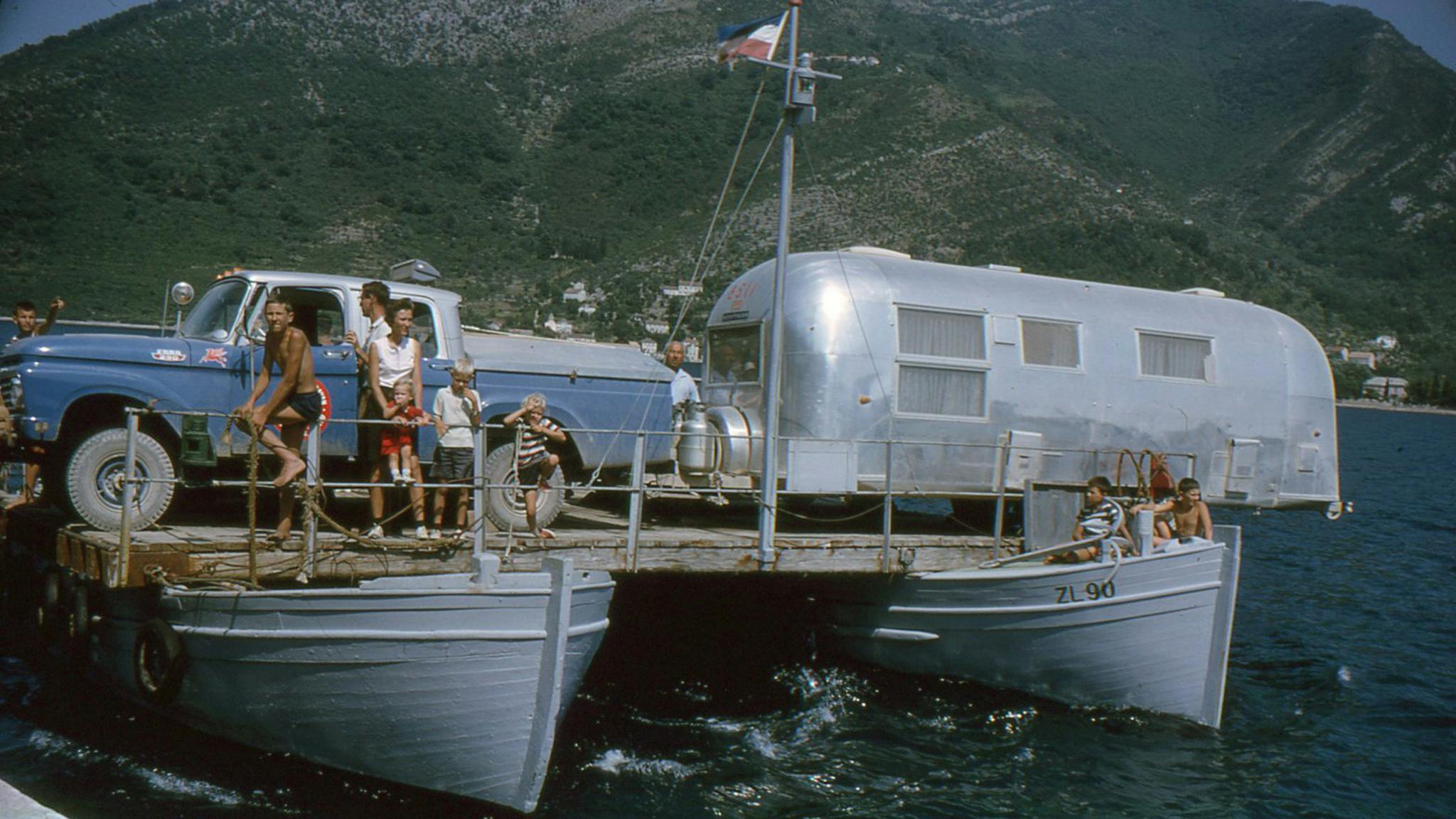 People on a boat that is carrying an Airstream travel trailer across the water during the Around the World Caravan.