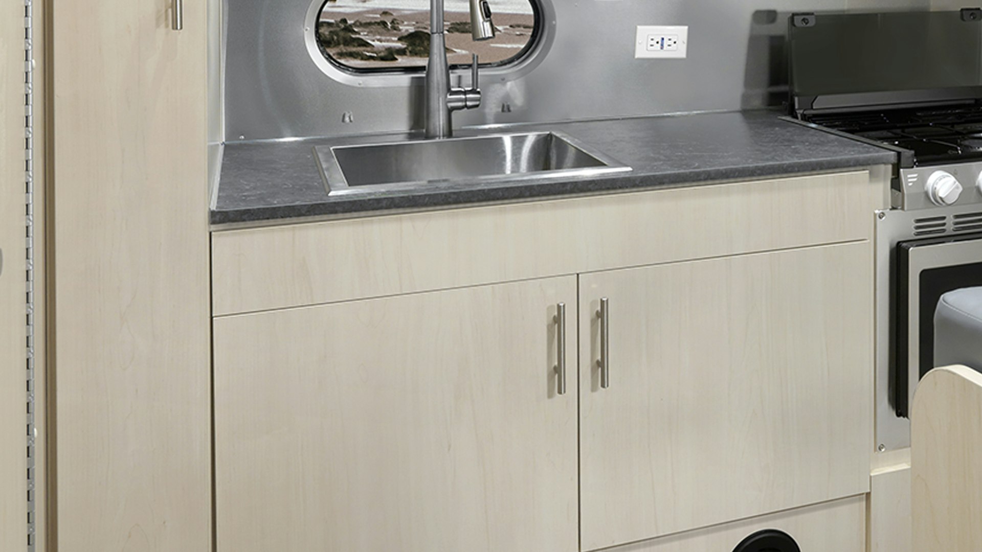 The cabinets below the kitchen sink in the galley of the Airstream Flying Cloud 23 twin travel trailer that is parked next to a body of water with rocks.