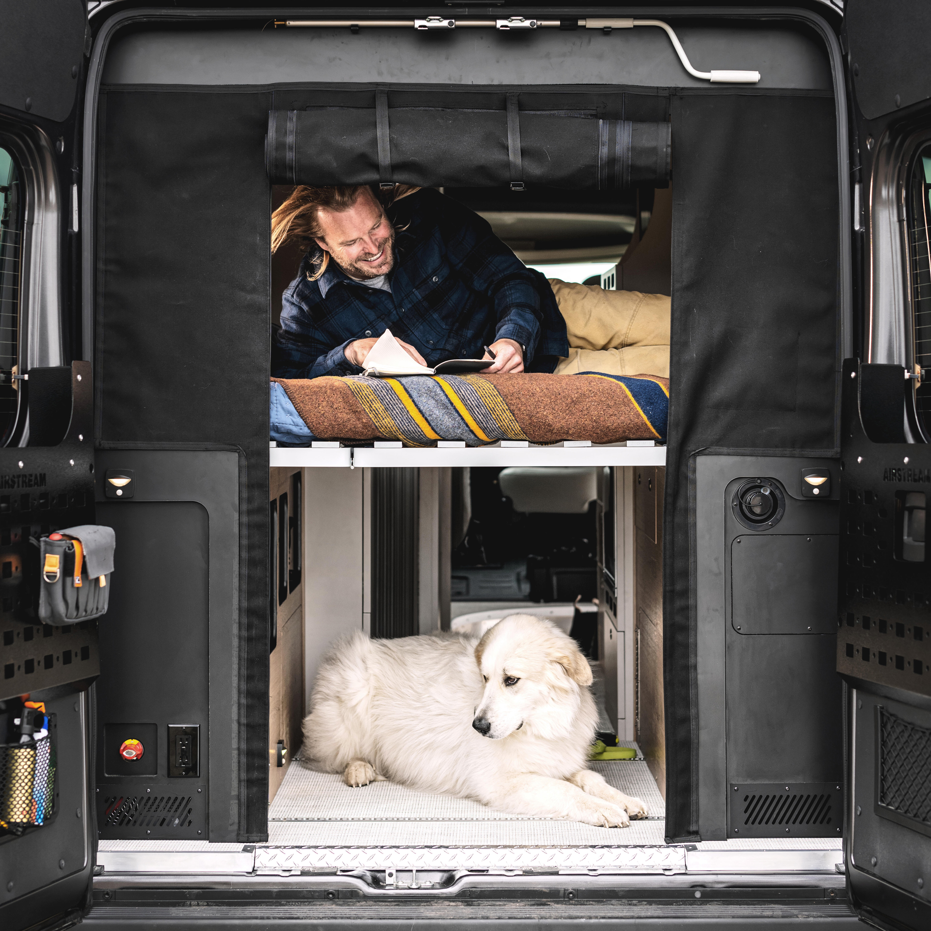 A guy laying in the bed of the Airstream Rangeline touring coach writing with the back doors open and his dog laying in the aisle way under him.