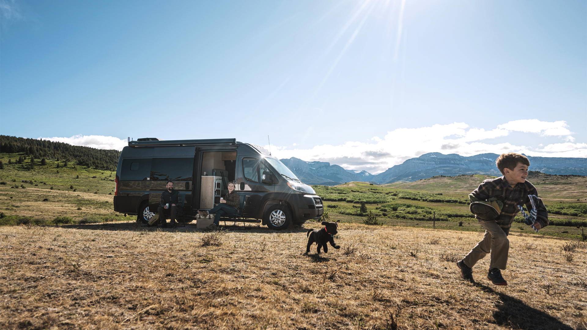 A boy and his puppy running while his parents sit outside under the awning of the Airstream Rangeline Touring Coach