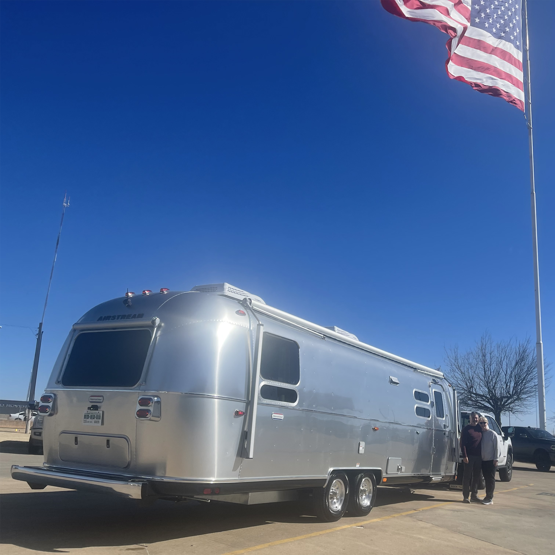 吉姆和苏珊微笑着在他们的新Airstream国际旅行拖车露营车旁拍照，美国国旗飘扬在蓝色的天空中。