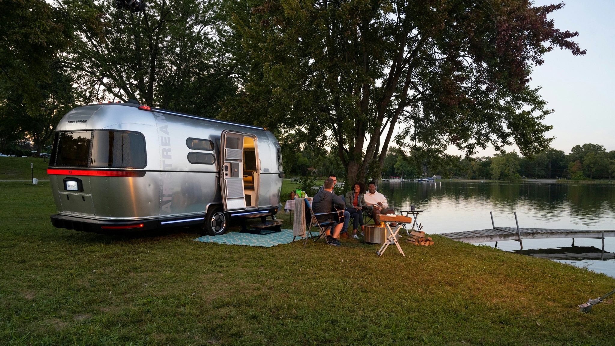 People sitting around a fire and by a lake and the Airstream all-electric Airstream travel trailer that is a concept vehicle.