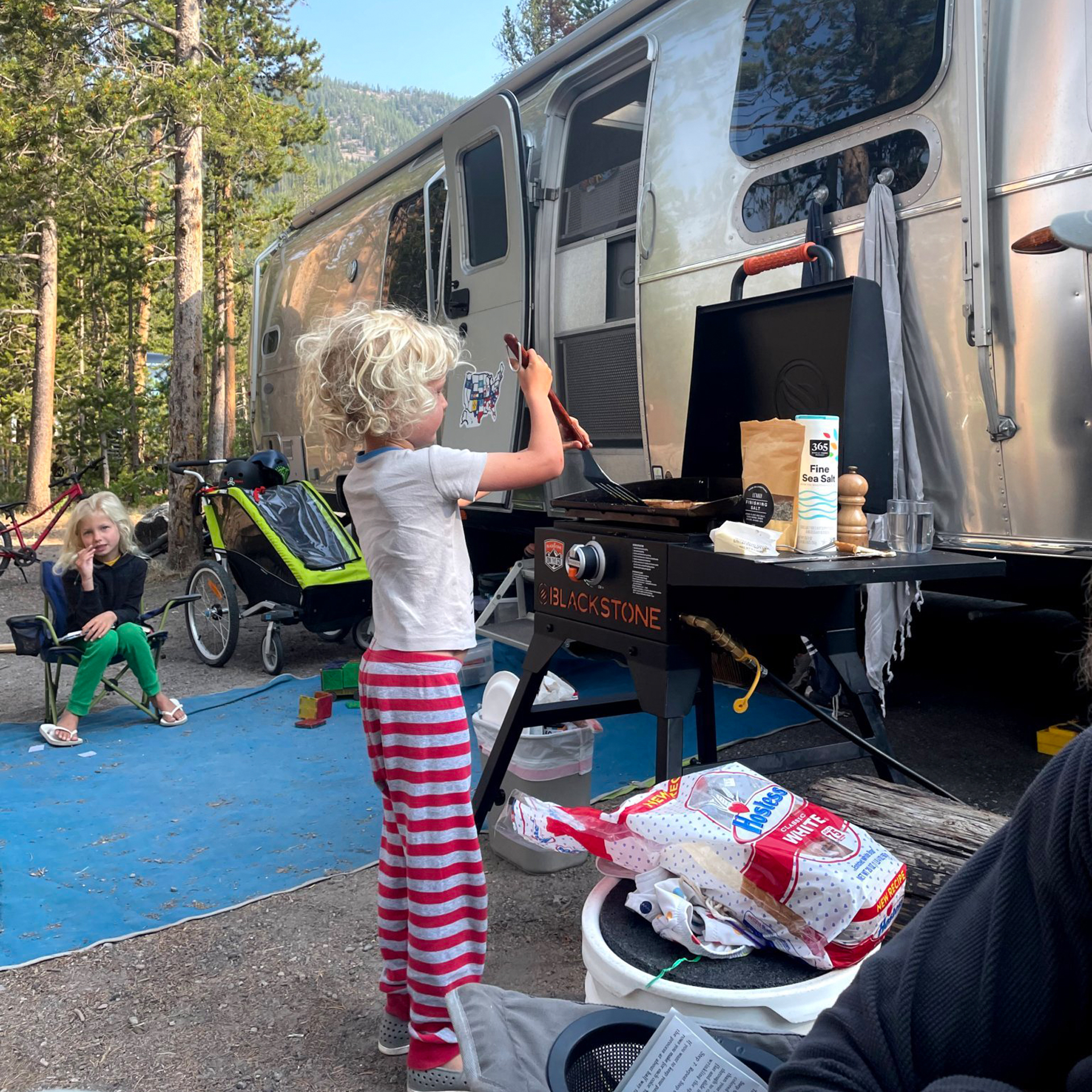 A little kid cooking french toast on a grill next to the Airstream travel trailer while a family camps.