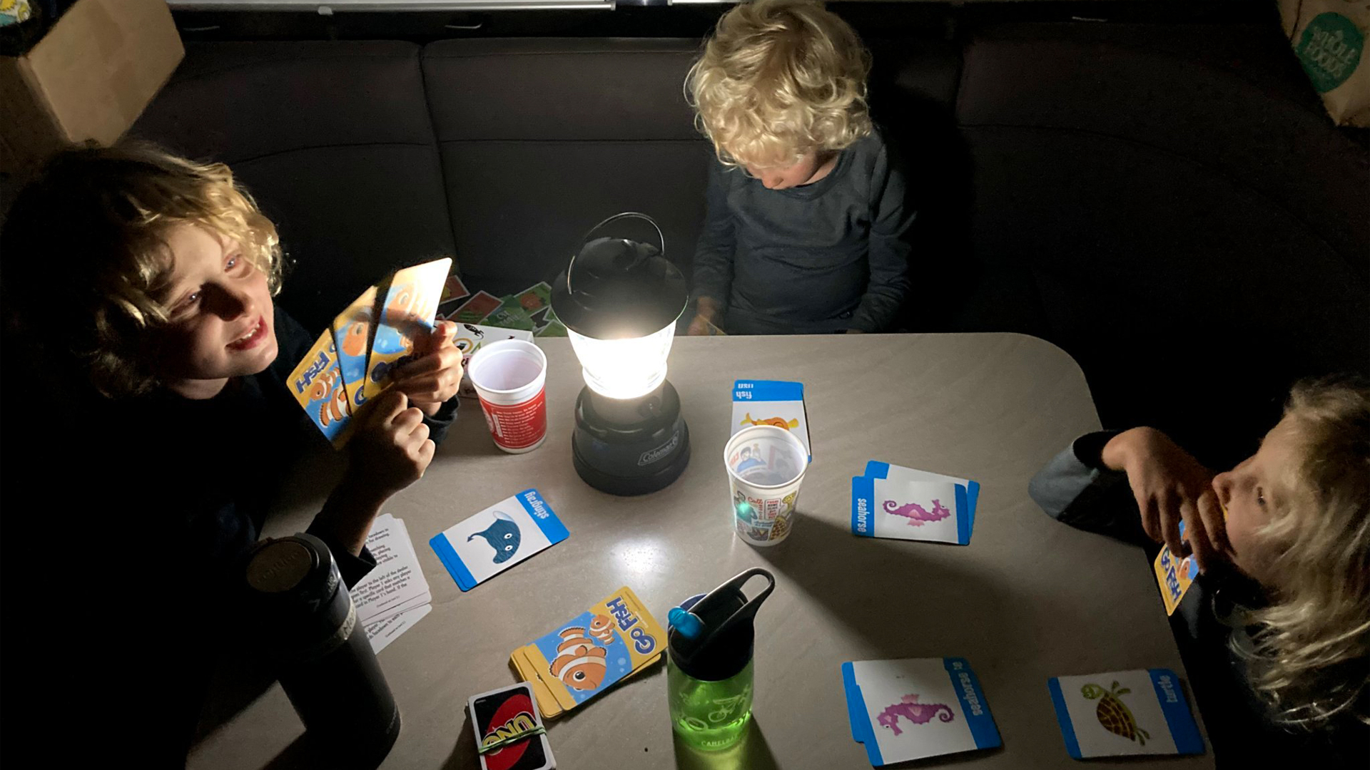 三个孩子坐在他们的Airstream旅行拖车的桌子旁，晚上开着夜灯玩钓鱼游戏。