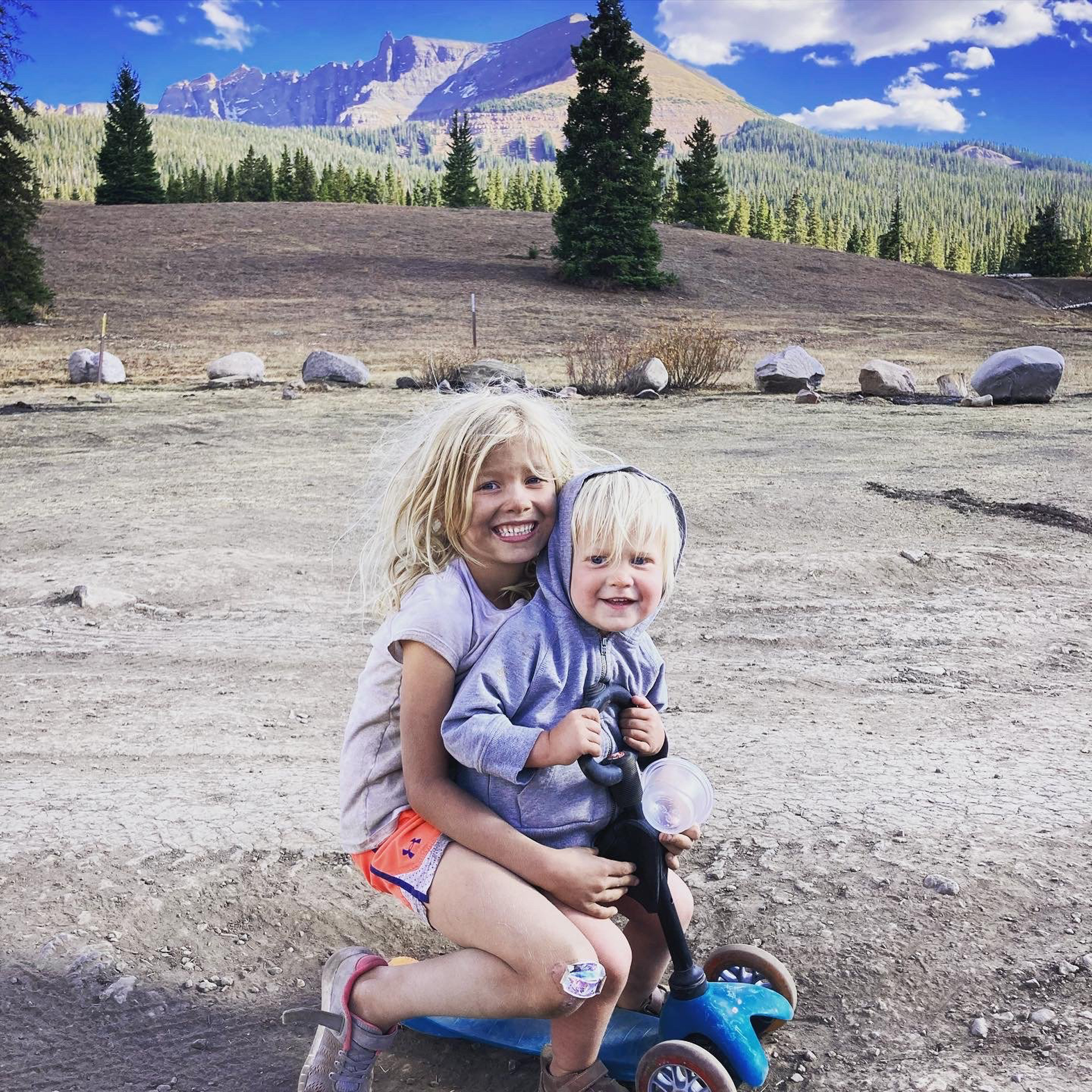 两个孩子骑着滑板车在泥土里玩，同时他们在他们的Airstream旅行拖车里露营
