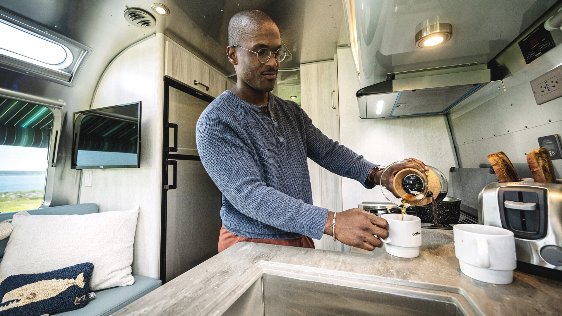 在他的Airstream国际旅行拖车的厨房里，一个男人把咖啡倒进他的咖啡杯里。