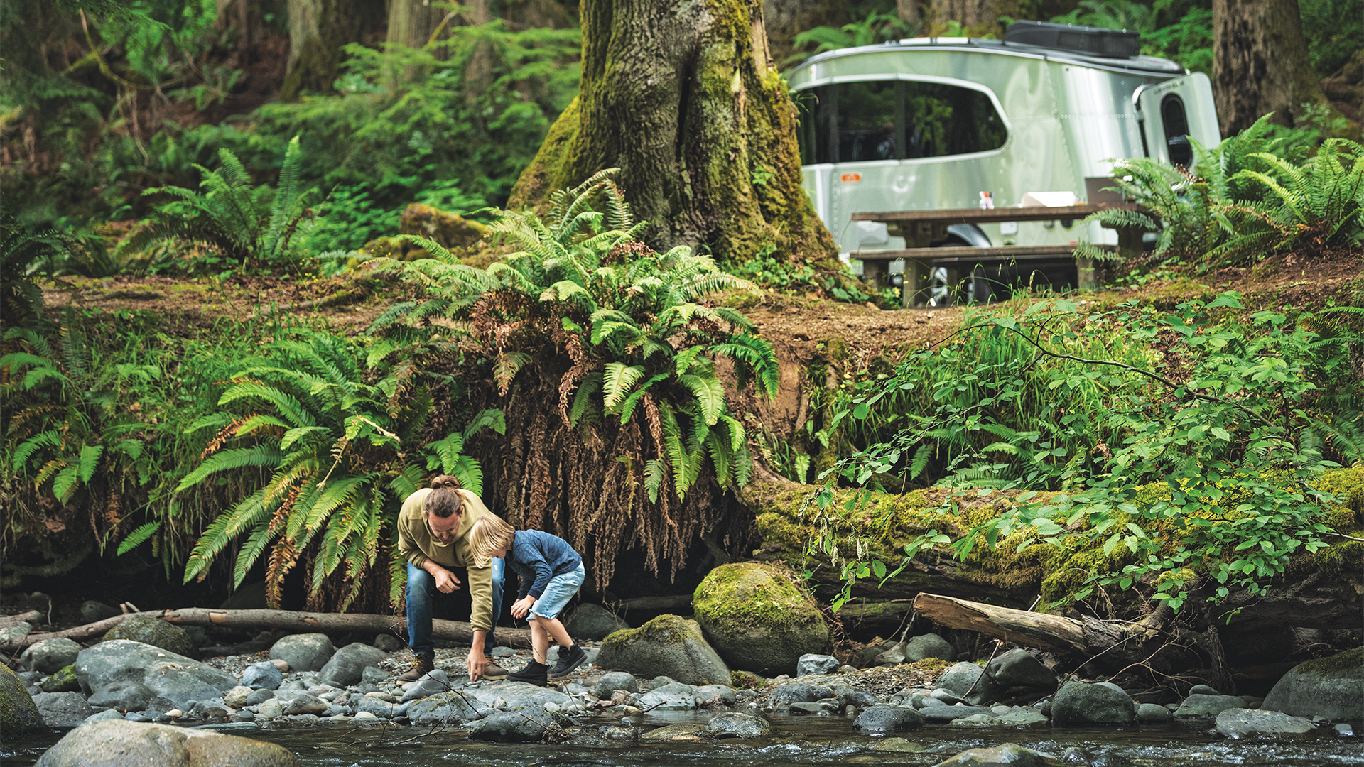 An Airstream Basecamp travel trailer parked in the mountains while a dad and boy are camping and playing in the river.