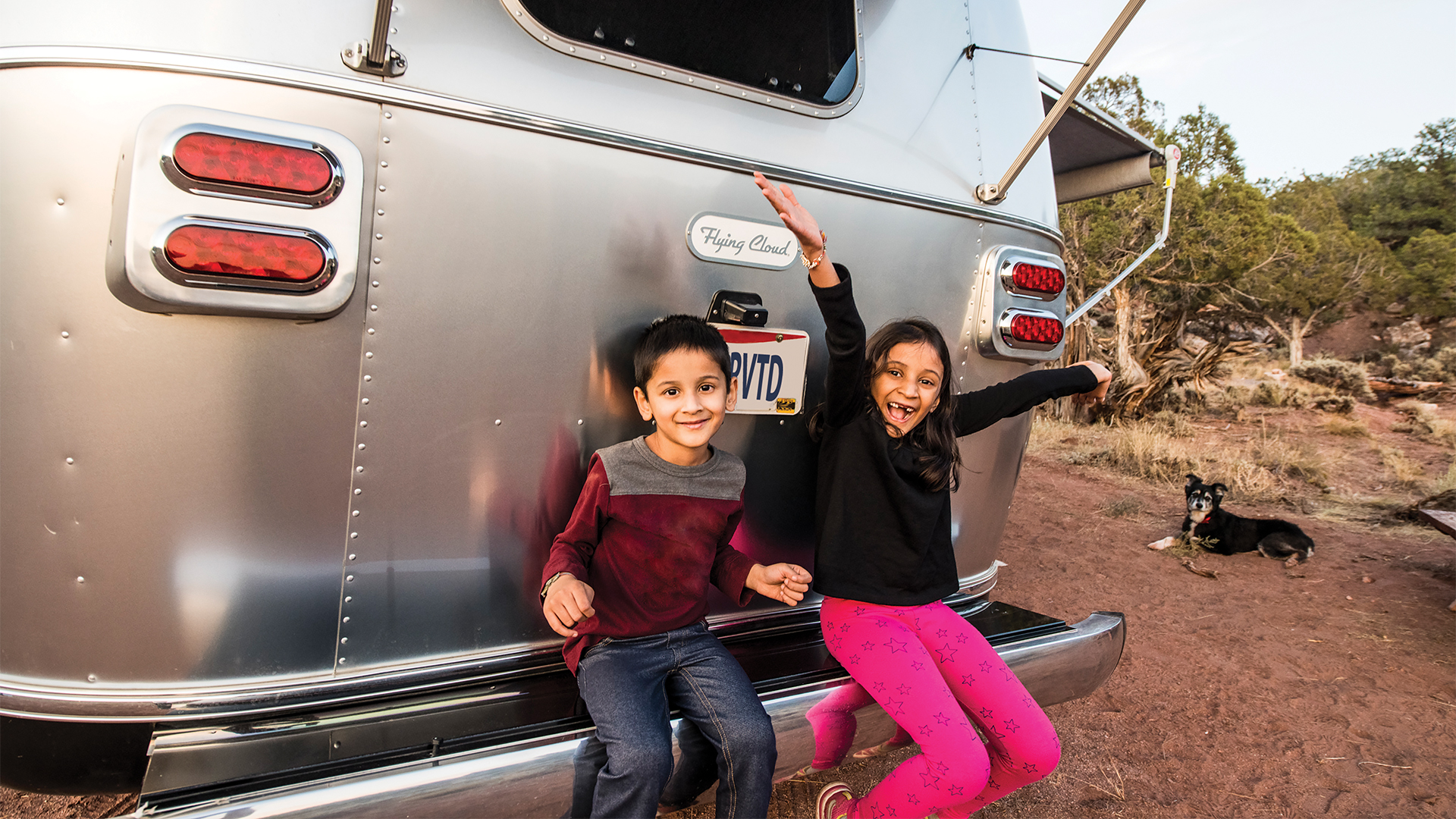 Two kids sitting on the bumper of their Airstream Flying Cloud travel trailer while they camp.