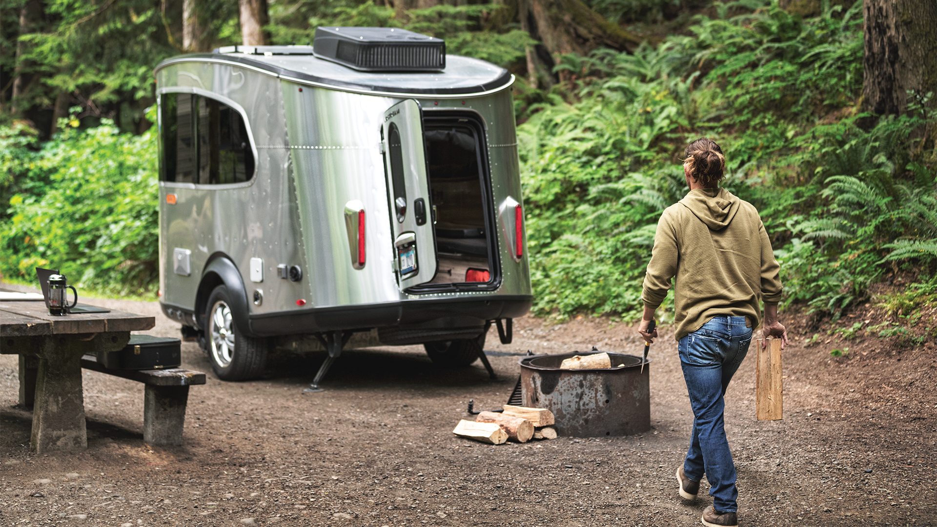 A man carrying a piece of firewood to a fire ring while he camps in a forest in his Airstream Basecamp travel trailer.