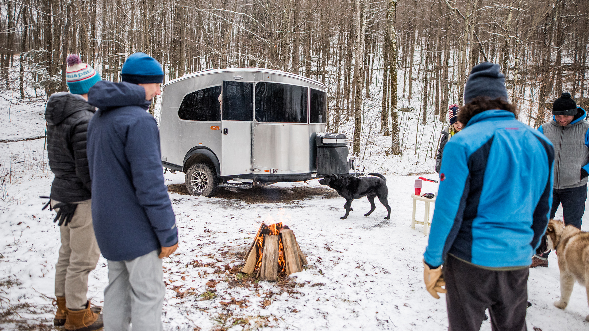 Camper friends stand around a campfire in the snow that is next to an Airstream Basecamp travel trailer.
