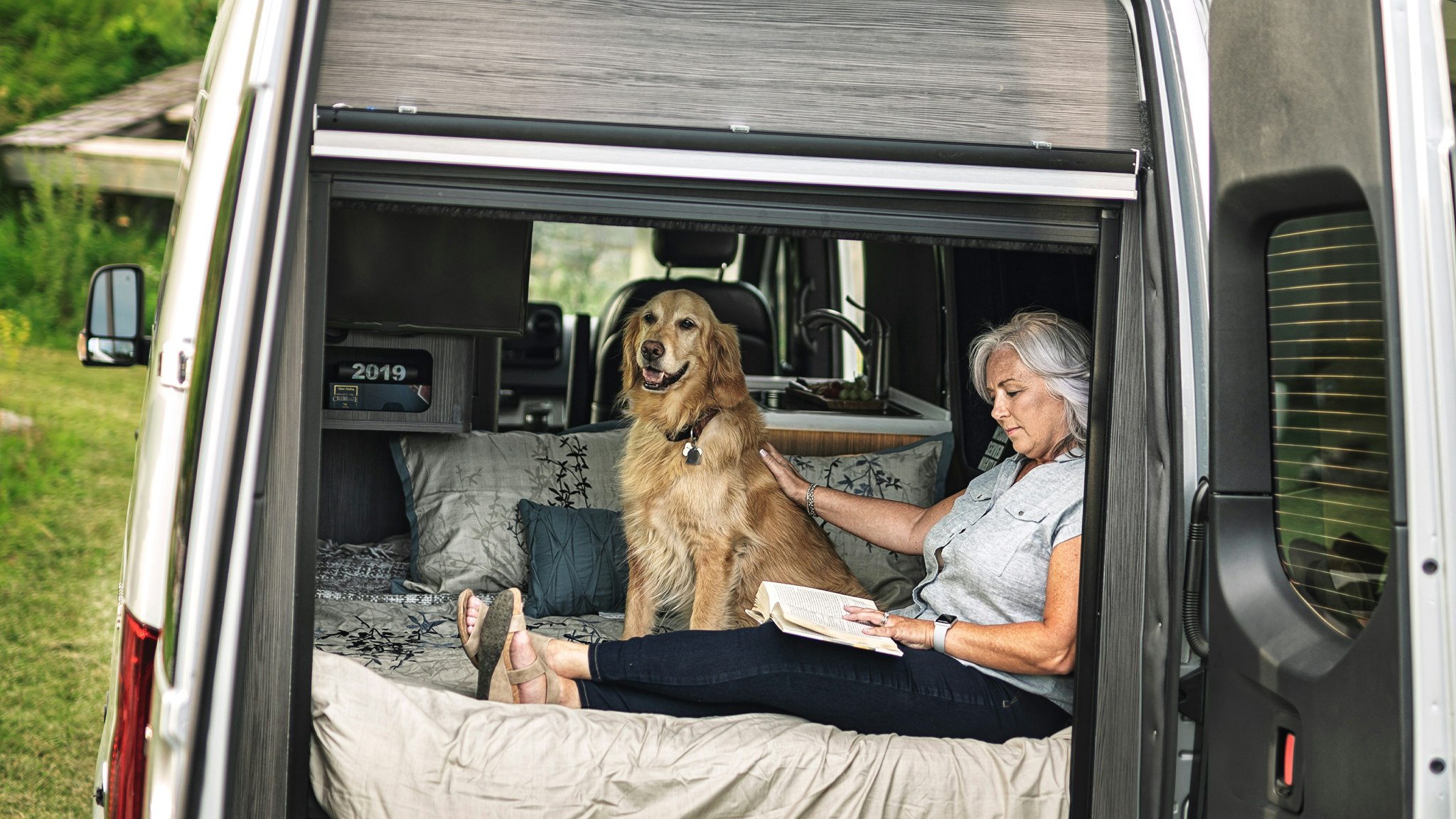 A woman and her dog sitting in the back of an Interstate 19 Class B motorhome while they sit in the bed while she reads with the rear doors open.