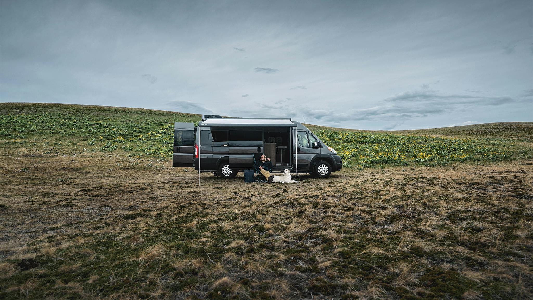 A man and his white dog sitting outside under the awning of their Airstream Rangeline Class B Motorhome that is parked in an open field.