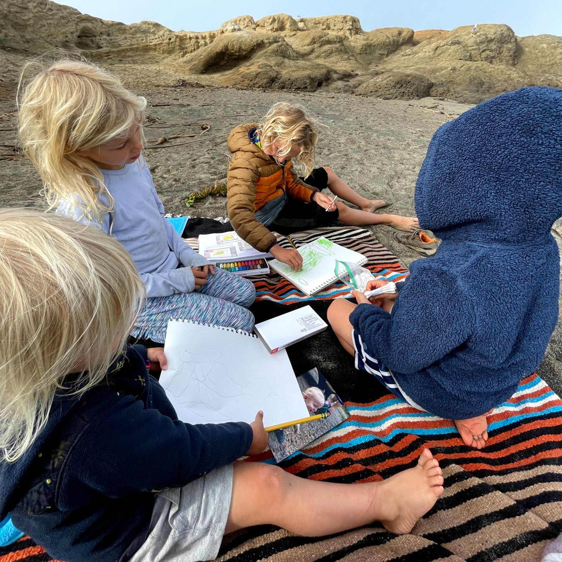Children sitting on a blanket in the sand coloring.