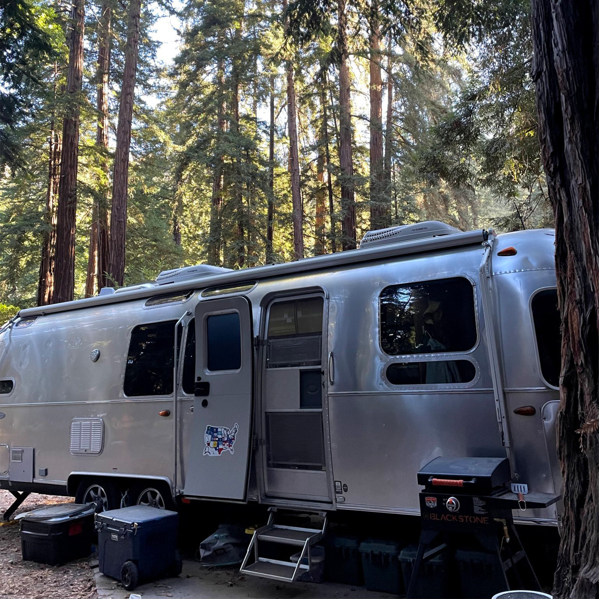 An Airstream travel trailer parked in a woods while a family camps.