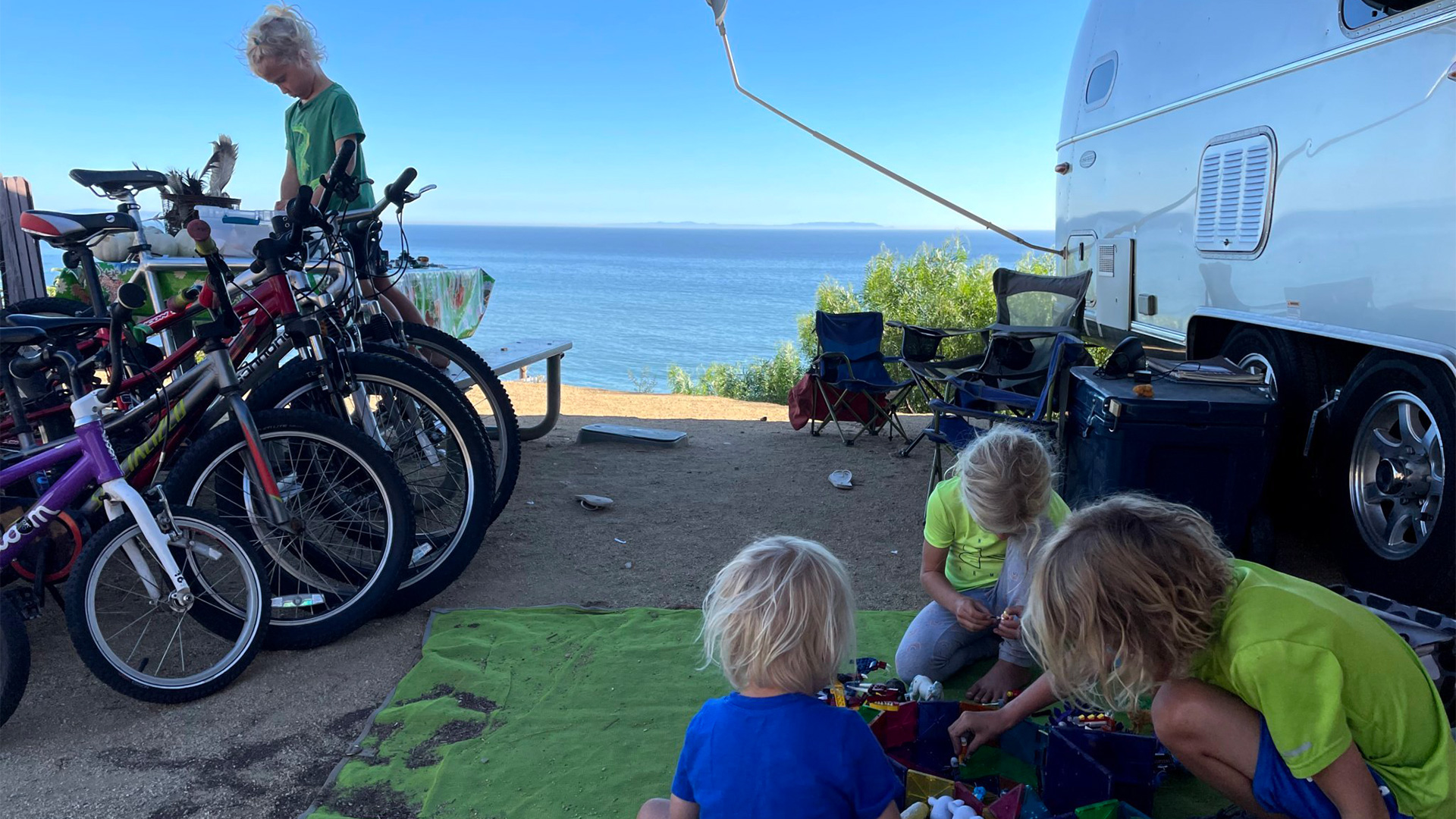 Three kids playing outside on a mat under the awning of an Airstream travel trailer while another camper gets a bike out.