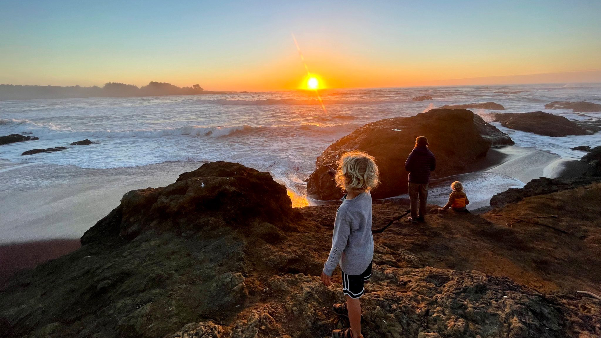 Kids and their parent standing on rocks by a body of water during a sunset.