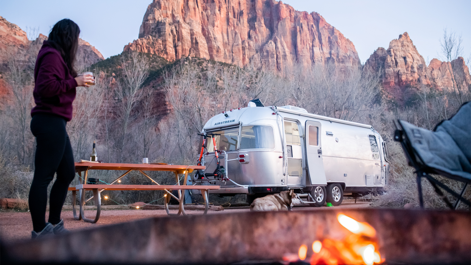 A woman carrying a cup to sit down by the campfire that is made while camping in her Airstream travel trailer in the desert.