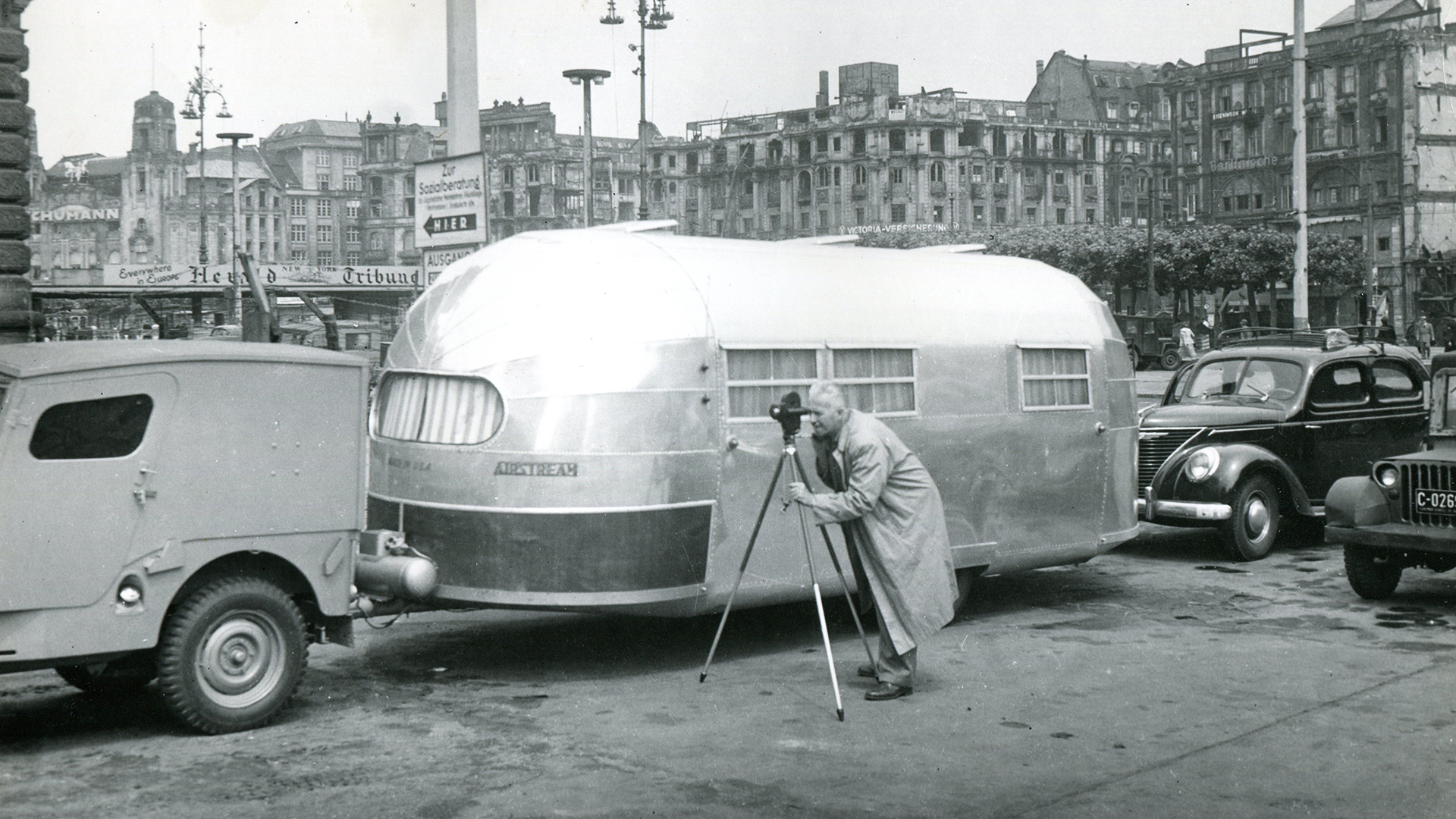 A vintage image of a man standing next to his Airstream travel trailer with a video camera on a tripod.