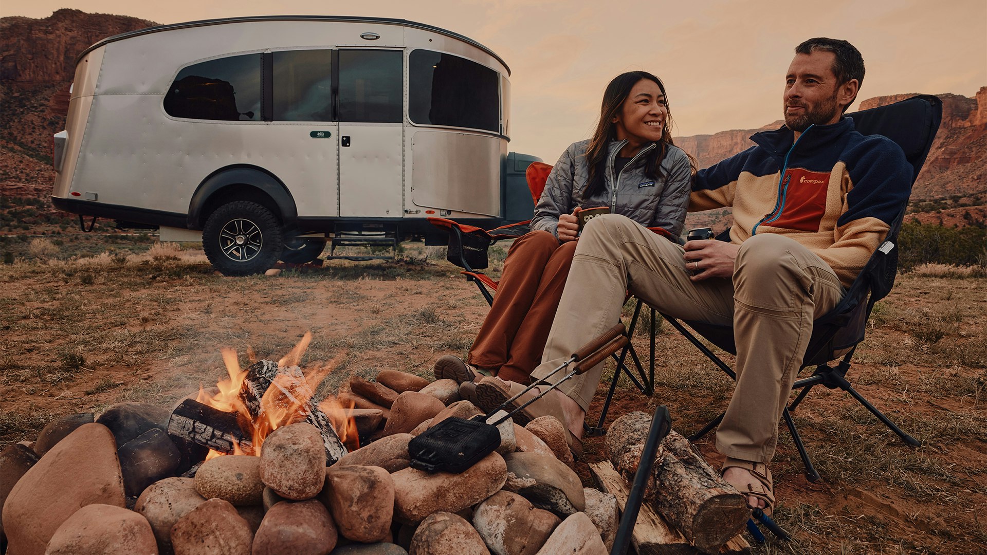 A man and woman camper sitting around a campfire outside of their Airstream REI Co-op Special Edition Basecamp 20X.