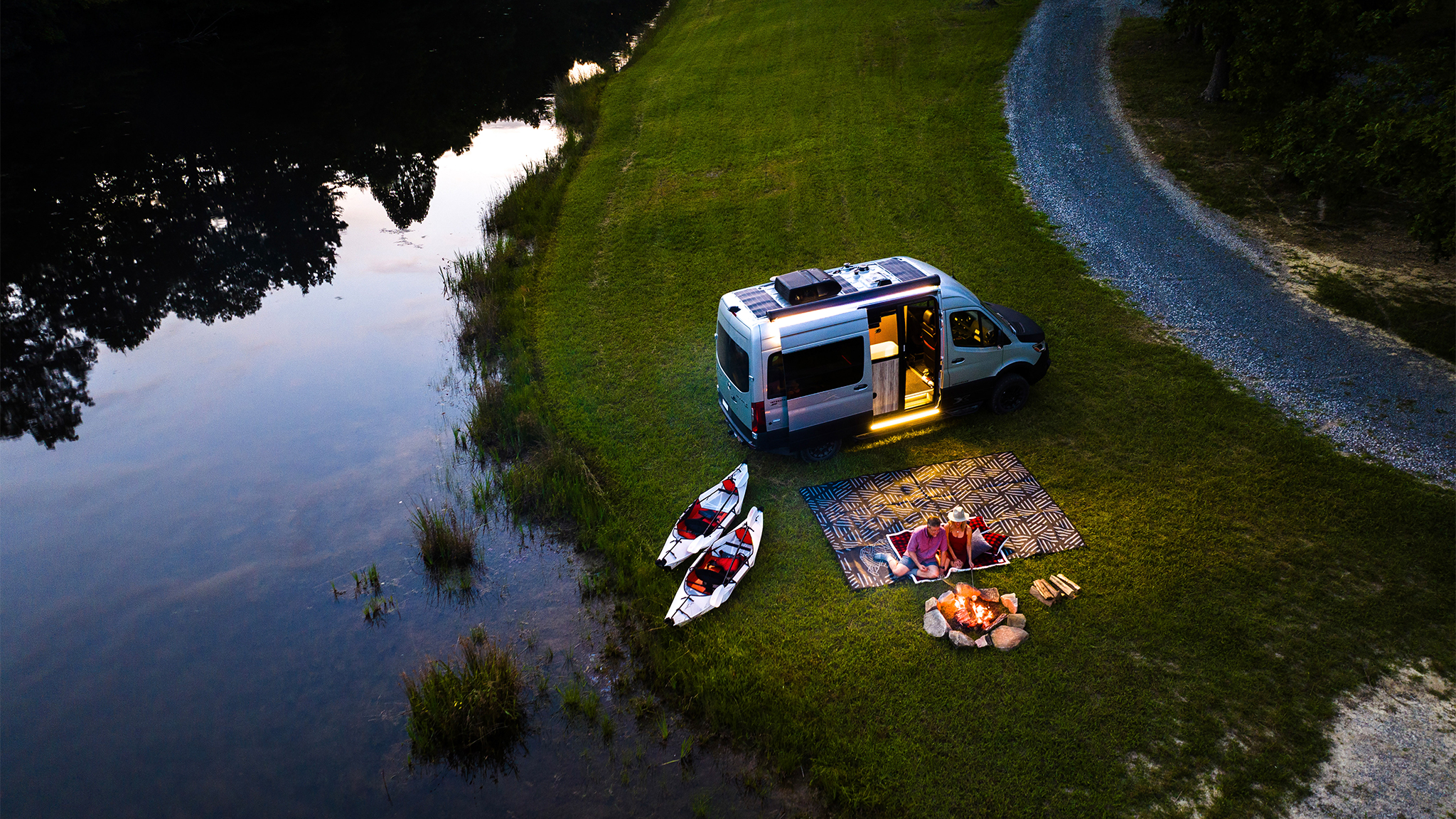 An ariel shot of the Airstream 19X Touring Coach parked next to a lake with a couple sitting by a campfire at night.
